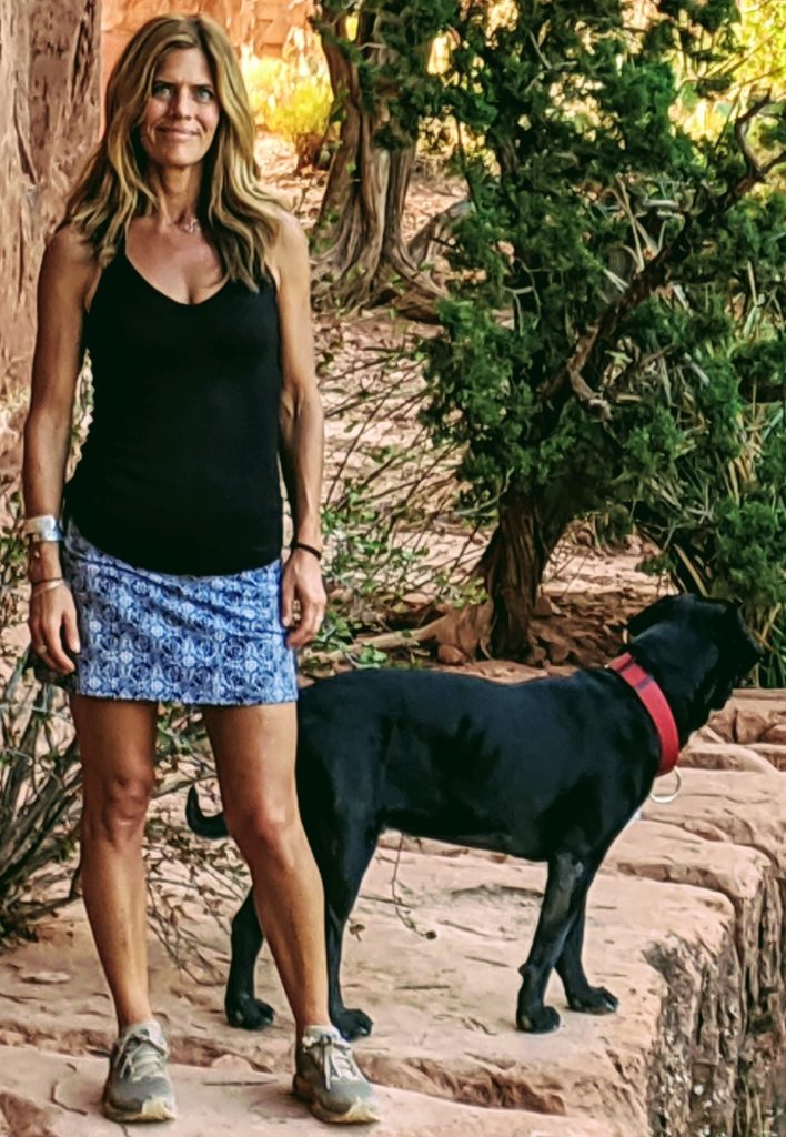 Britta Nelson, a certified yoga instructor at Full Circle Yoga & Therapy in Salt Lake City is pictured outdoors with her dog. Britta is a caucasian female with long, wavy hair that is blondish brown. She is muscluar in stature and pictured wearing a black tank top and blue patterned skirt with tennis shoes. Her dog is a black lab.