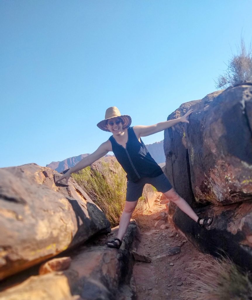 Telesa Nichols, certified yoga instructor at Full Circle Yoga & Therapy. Telesa is pictured outdoors in a red rock desert area on a sunny day. She is wearing her hair tucked into a straw hat, sunglesses, black sleeveless shirt, and gray knee-length shorts.