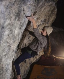 Raleigh Ramos is pictured bouldering during the evening. Raleigh has long, wavy dark hair and is wearing navy athletic pants, a brown heather sweater, and a cream colored beanie hat.