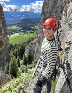 Samantha Curtis is pictured mountain climbing in sunny weather. She is wearing a red helmet, a black and white striped, long sleeved shirt, and traditional hiking/climbing harnesses and gear.