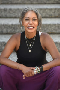 Professional photo of Nikki Myers, founder of Yoga of 12 Step Recovery. Nikki is an African American woman pictured seated on grey granite steps. Her naturally grey hair is loosely pulled back away from her face. She is smiling and wearing a black tank top and dark purple pants. 
