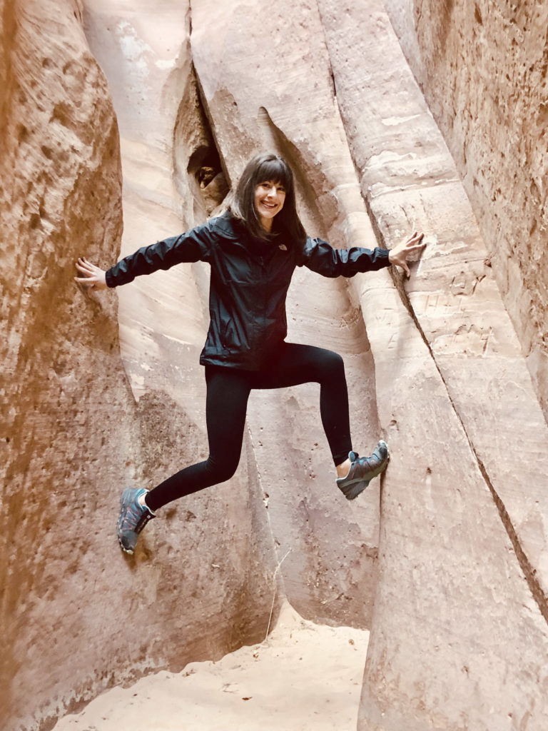 Jennifer Rohn, Director of Operations of Full Circle Yoga & Therapy is pictured balancing between rock walls of a redrock slot canyon in Southern Utah. She has straight, shoulder-length brown hair with bangs and is wearing a black windbreaker jacket, black yoga leggings, and tealish green train shoes.